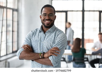 手を組んでポーズをとる幸せなアフリカ系アメリカ人の中小企業の経営者のポートレート。ミレニアル世代の黒人男性チーム リーダーが微笑み、カメラを見て、後ろの近代的なオフィスで働く従業員。ヘッド・ショット