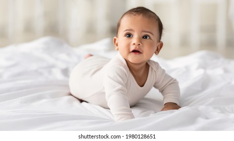育児のコンセプト。自宅で白いシーツの上に横たわるボディスーツを着たかわいいアフリカ系アメリカ人の赤ちゃんのポートレート。寝室のベッドの上を這う黒人の幼児。選択と集中、無料コピー スペース