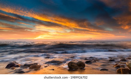 A Panorama Of A Colorful Ocean Sunset Sky As A With As A Gentle Wave Rolls In Among The Seashore beach Rocks And Sand