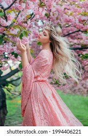 Fantasiefrauenprinzessin geht im blühenden Frühlingsgarten spazieren. hand berührt rosa blumen sakura-baum sommernatur. langes Spitzenkleid mit weiten Ärmeln. Blondes Haar fliegt im Wind. Elf Diadem-Tiara-Krone auf dem Kopf