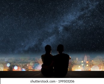 Silhouette of young Asian couple sit on wooden ground above the city under stars.