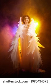 Una chica morena con un vestido elegante y alas de ángel blancas sobre un fondo negro. Modelo, actriz o bailarina posando en el estudio.