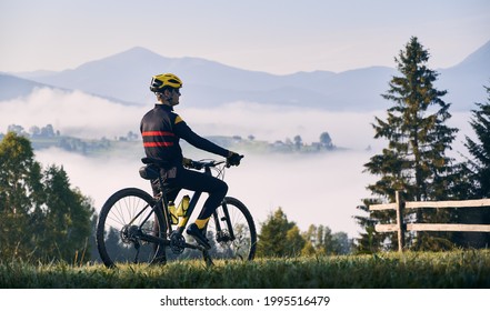 芝生の丘の上に自転車で立って、美しい霧深い山々を見ているサイクリスト。自転車に乗って雄大な山々の景色を楽しむ男性の自転車乗り。スポーツ、自転車、自然の概念。