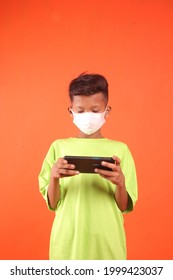 Profile shot of tan teenager standing and using his smartphone and wearing a mask and an oversized light green t-shirt, isolated on an orange background