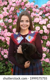 Halloween en Rusia. Chica de moda rusa, mujer en jardín de rosas con gato negro, animal, mascota. Mira con cuello bordado. Fiesta de Halloween. Estilo callejero. Florecer. Rosas rojas, flores. Traje de halloween