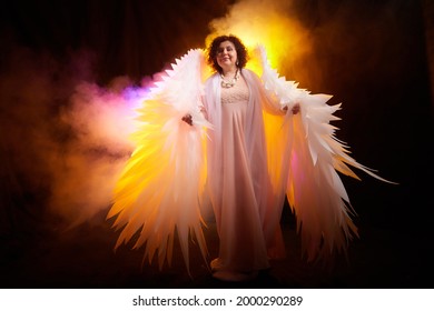 Una chica morena con un vestido elegante y alas de ángel blancas sobre un fondo negro. Modelo, actriz o bailarina posando en el estudio.