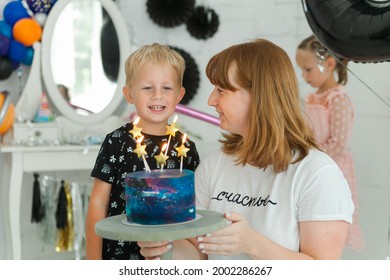 een vrolijke schattige lachende jongen in een zwart t-shirt viert zijn vijfde verjaardag thuis, versierd in ruimtethema. hij blaast kaarsjes op de taart met mama en doet een wens. "Geluk" op moeders t-shirt