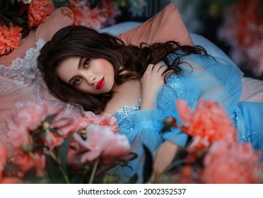 Portrait fantasy woman sleeping beauty lies on comfortable bed soft pillow. Background mystical garden, pink peonies flowers green trees. Fairy-tale girl princess cute face red lips makeup, blue dress