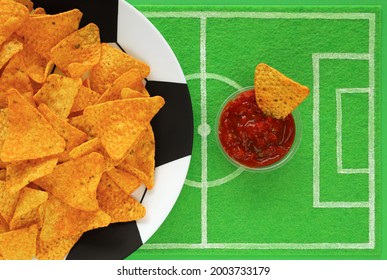 Football soccer party table. Corn nacho chips on big plate painted like soccer ball and one in red salsa sauce on dish mat like football field made of green felt, top view. Watch match broadcast.