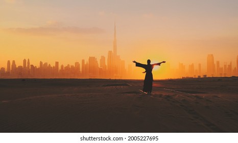 Arabische vrouw, gekleed in traditionele kleding van de VAE - abayain die haar handen opsteekt op de zonsondergang in een woestijn met het stadssilhouet van Dubai op de achtergrond.