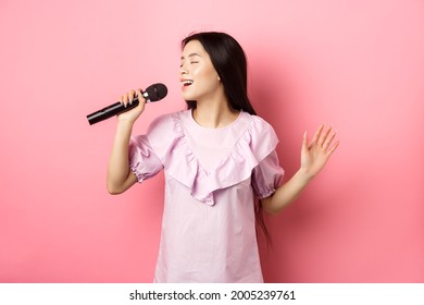 美しいアジアの女の子は歌を歌い、マイクで歌い、ロマンチックな笑顔を浮かべ、ピンクの背景にドレスを着て立っています