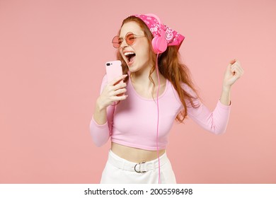 若い歌手女性 20 代バラの服を着てバンダナ メガネ ヘッドフォン カラオケ クラブでマイクで歌を歌ってパステル ピンクの背景のスタジオ ポートレートに分離されたモバイル携帯電話で声を録音します。