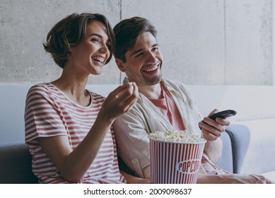 Side view young cheerful couple two friend woman man 20s wearing casual clothes sit on sofa hold takeaway bucket eat popcron watch movie rest indoors at home flat together. People lifestyle concept.