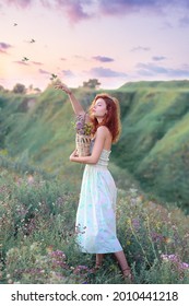 A beautiful red-haired girl raising her hand up towards the birds against the background of a forest landscape and a stunning pink-lilac sunset, in a light summer sundress with a basket of wildflowers