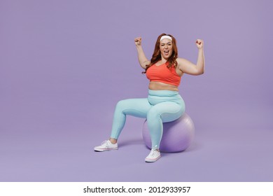 Full length side view young chubby overweight plus size big fat fit woman in red top warm up train sit on fit ball do winner gesture isolated on purple background gym. Workout sport motivation concept