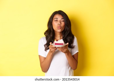 Portrait of beautiful african-american girl celebrating birthday, blowing lit candle on b-day cake and making a wish, standing over yellow background