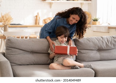 Un niño emocionado que recibe un regalo de cumpleaños de mamá sentada en un sofá en la sala de estar. Concepto de celebración de vacaciones para niños con una madre amorosa dando un regalo a un adorable hijo de preescolar felicitando al niño