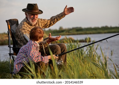 Grootvader en jongetje vissen samen 's avonds bij zonsondergang in de zomerdag op rivier op het platteland, zijaanzicht op blanke familie van meerdere generaties, oudere man leert kindjongen vissen