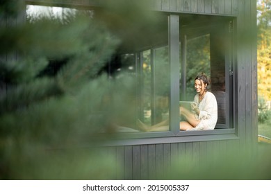 Junge Frau, die sich in einem schönen Landhaus oder Hotel ausruht, mit einem Tablet auf der Fensterbank sitzt und die schöne Aussicht auf den Kiefernwald genießt. Ansicht von außen. Schöne Urlaubsziele