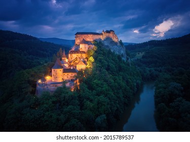 Letecký, dramatický pohľad na Oravský hrad, fantazijný hrad na vysokej skale nad riekou Orava, osvetlený oranžovými lampami na tmavom, zamračenom pozadí. Miesto, ktoré používajú tvorcovia filmov. Slovensko.