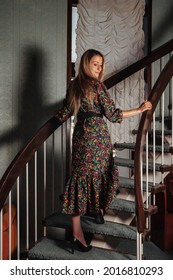 Retrato mujer adulta actriz posando vestida con ropa de estilo retro en la escalera de casa en la sala de estar vintage. Hembra en vestido de color de estilo antiguo en flores. Concepto de moda retro y moda de tendencia obsoleta