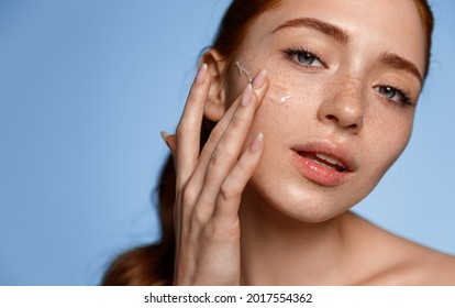 Protección de la piel. Primer plano de una joven pelirroja tocándose la cara para aplicar crema hidratante, fondo azul