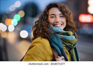 Potret wanita bahagia di jalan kota melihat ke kamera. Gadis cantik berdiri di luar ruangan di malam musim dingin dengan senyum lebar. Mahasiswa ceria mengenakan mantel dengan syal wol saat senja.