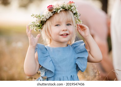 klein meisje draagt ​​een bloemenkrans op haar hoofd in een veld op zonnige zomerdag. baby in een blauwe jurk. Portret van schattig klein kind buitenshuis. fijne vakantie jeugd.