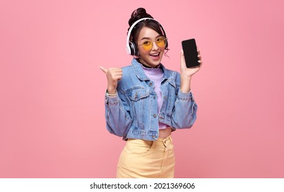 Ung asiatisk teenagekvinde, der viser smart telefon, hun lytter til musik i hovedtelefoner isoleret på lyserød baggrund.
