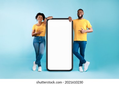 Great mobile app. African American couple pointing at giant smartphone with mockup, promoting application or website, advertising product or service, blue background. Full length