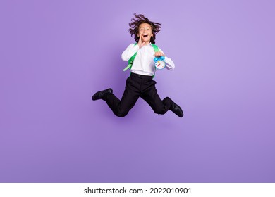 紫色の背景に分離された印象的な小さな黒髪の少年ジャンプ ホールド時計着用バッグ シャツ ズボン スニーカーのフル サイズの写真