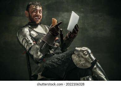 Watching funny video. Portrait of one bearded man, medeival warrior sitting with tablet eating hot dog over dark background. Combination of medeival and modern styles. Copyspace for ad