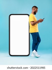 Full length of young Afro guy using cellphone while leaning on giant mobile phone with empty white screen, blue studio background. Mockup for app or website, space for advertisement