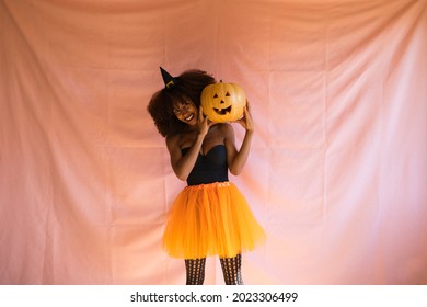 young african american woman dressed as a witch for halloween party and holding a pumpkin in her hands. She wears a witch's hat and an orange skirt. The woman smiles at the camera with the pumpkin.