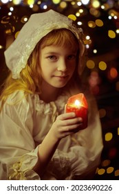 Cute little girl in a nightgown and a cap looks at the candle on Christmas night. Magic time. Christmas in the old days. 