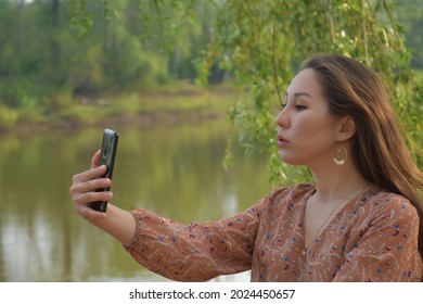 Side view of young pretty Asian female with long dark brown hair in dress taking selfie in park immersed in greenery in daytime on blurred background 