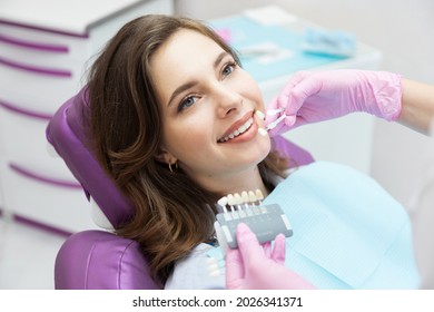 Dentista que elige el color del tono del esmalte de los dientes para una paciente joven y sonriente antes del procedimiento de blanqueamiento. Mujer feliz con hermosa sonrisa blanca en cita estomatológica. Odontología y estomatología.