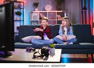 Hübscher Bruder und Schwester, die mit gekreuzten Beinen auf einer bequemen Couch sitzen und fernsehen. Glückliche zwei Kinder in Freizeitkleidung, die zu Hause lustige Cartoons oder interessante Filme genießen.