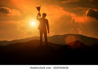 Silueta de un hombre de negocios sosteniendo un trofeo en la cima de una montaña con la puesta de sol. El concepto de un negocio exitoso o un gran ejecutivo para llevar a la organización al éxito.