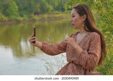 Side view of young pretty Asian female with long dark brown hair in dress taking selfie in park immersed in greenery in daytime on blurred background