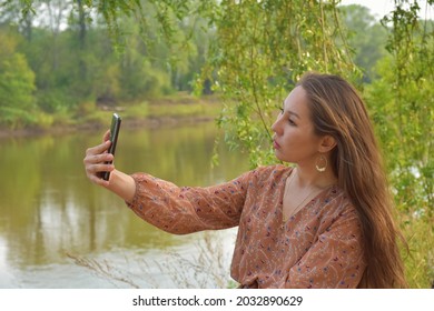 Side view of young pretty Asian female with long dark brown hair in dress taking selfie in park immersed in greenery in daytime on blurred background 