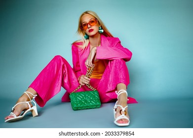 トレンディなピンクのフクシア色のスーツを着たファッショナブルな女性で、クラシックなブレザー、幅広のズボン、オレンジ色のメガネ、ストラップ付きのサンダル、緑のキルティング バッグを持っています。全身ファッション ポートレート。コピースペース
