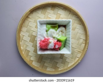 Lokaal Maleisië traditioneel dessert. Lompat tikam of springsteek in vierkante keramische kom. Ronde bamboe dienblad achtergrond. Gelei met pandan-smaak, roze kleefrijst, kokosmelk en palmsuiker. augustus 2021