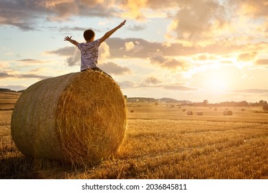 Boy sitting on a hay bale with arms raised in summer watching the sunset concept for worship, praise, religion or carefree childhood
