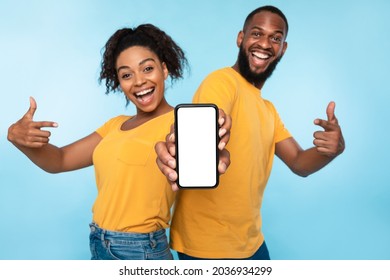 Anuncio de aplicación móvil. Una pareja afro emocionada señala un teléfono celular con pantalla en blanco y recomienda una nueva aplicación sobre un fondo de estudio azul, enfoque selectivo. Maqueta para sitio web
