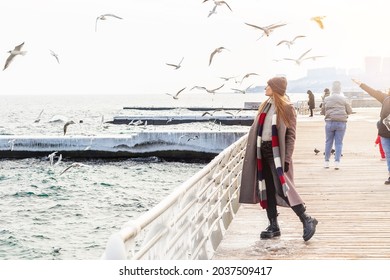 Gehende Frau auf dem Pier im Winter. Stilvolle Dame mit Wintermantel, groben Stiefeln, Schal und Hut, die allein am Meer steht