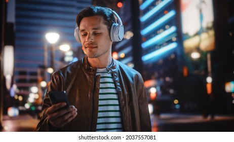 Portrait of Handsome Man Wearing Headphones Walking Through Night City Street Full of Neon Light. Smiling Stylish Man Listening to Music, Enjoying Podcast, Talk Show.