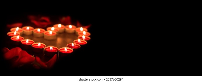 Kerzen brennen im Dunkeln. Rote Kerzen sind in Form eines Herzens auf schwarzem Hintergrund angeordnet. Rosenblätter sind um die Kerzen verstreut. Banner mit Platz für Text. Urlaubskarte.