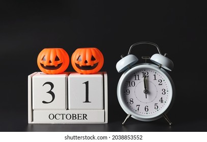 Front view of white vintage alarm clock show 12 o'clock  and wooden calendar October 31 with  plastic halloween pumpkins on black background. Halloween party concept.