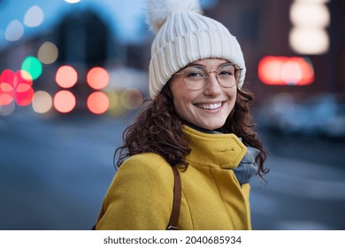 Potret wanita muda yang bahagia mengenakan topi hangat dengan kacamata di jalan kota dan melihat ke kamera. Murid cantik dengan topi dan mantel musim dingin berjalan di jalan kota saat senja dengan ruang fotokopi.